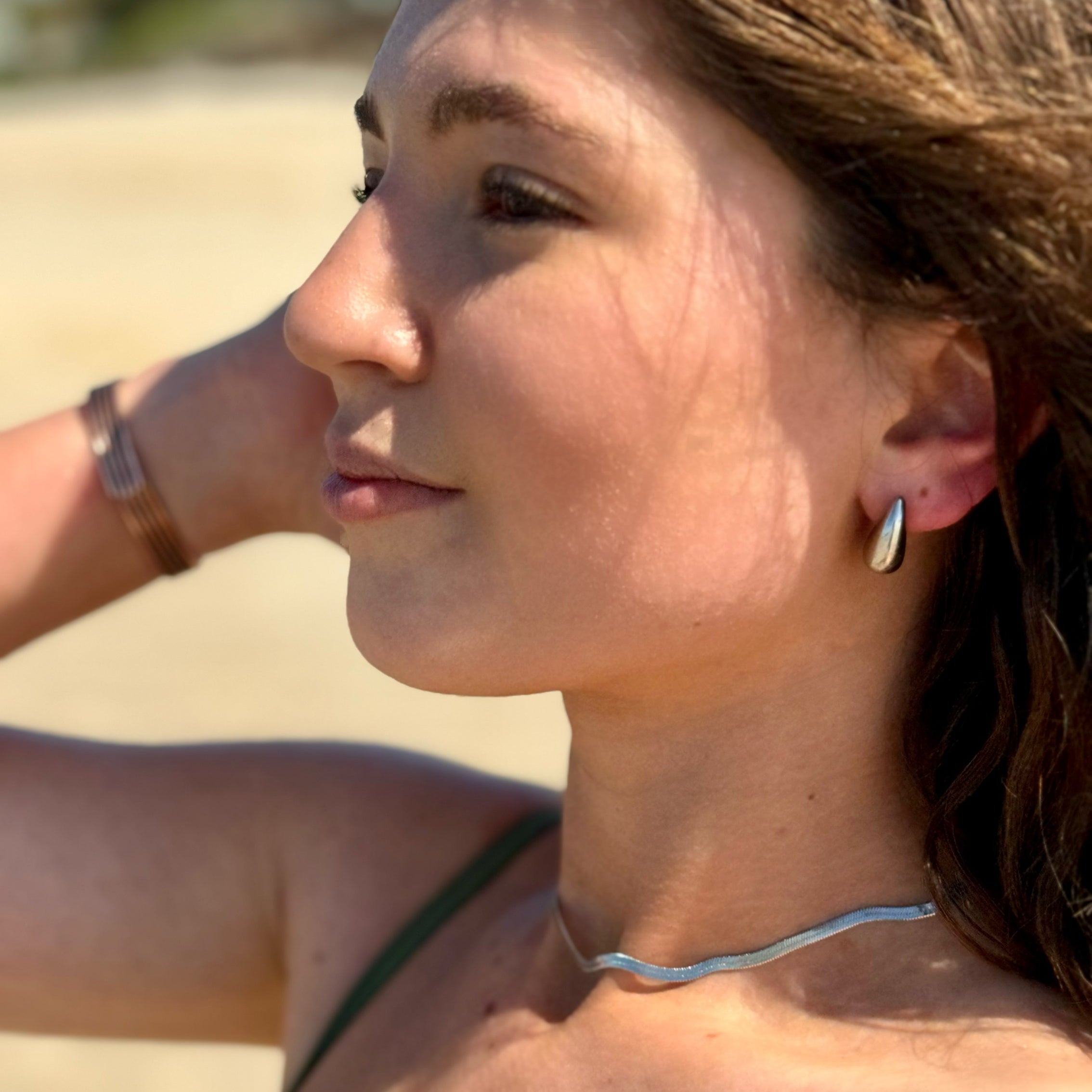 A woman in profile at a beach wearing a thin silver wavy necklace and a small silver hoop earring, with a green bikini top visible against a sandy background.